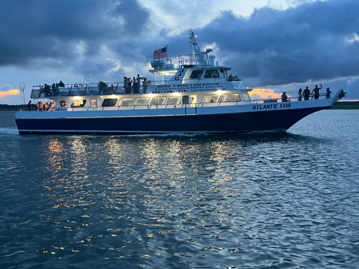 Large boat with lights on water at dusk with cloudy sky.